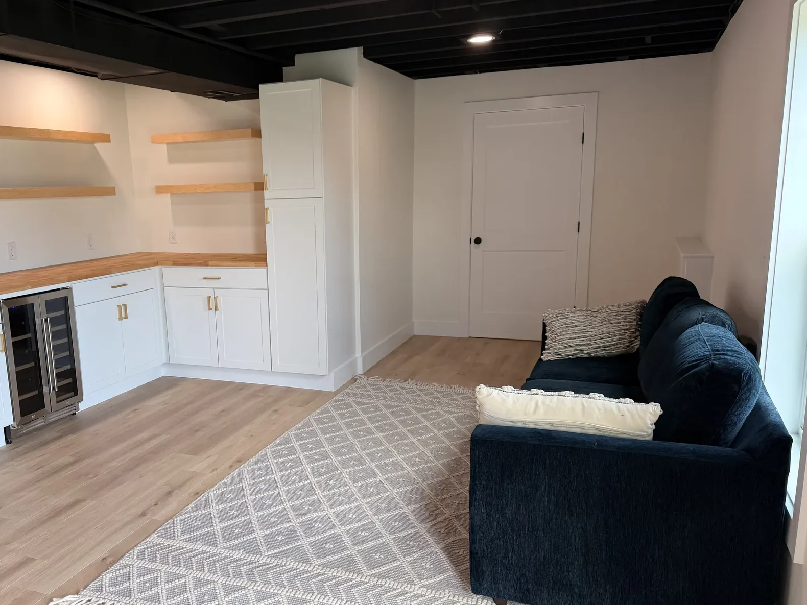 After: Finished basement with white shaker wet bar, butcher-block counter, floating shelves, beverage fridge, navy sofa and rug