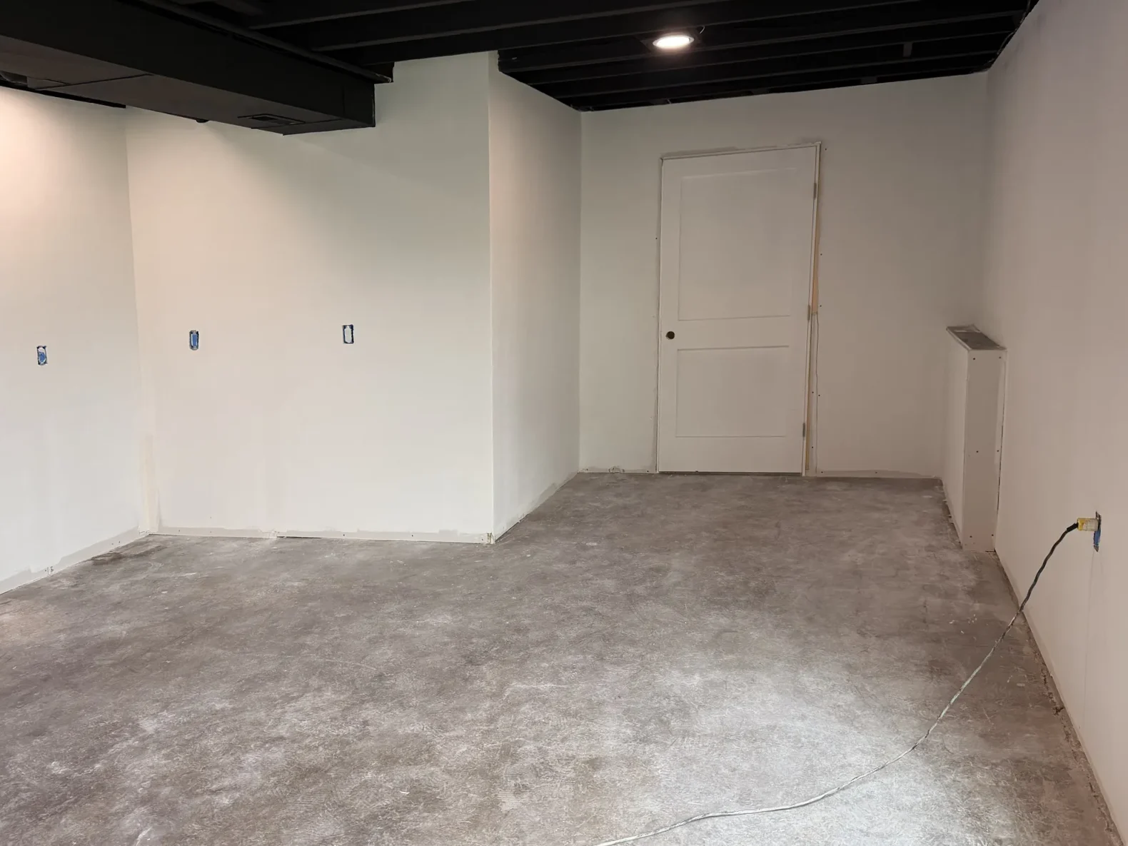Before: Basement corner with white painted walls, electrical boxes where cabinets will go, a new interior door and bare concrete