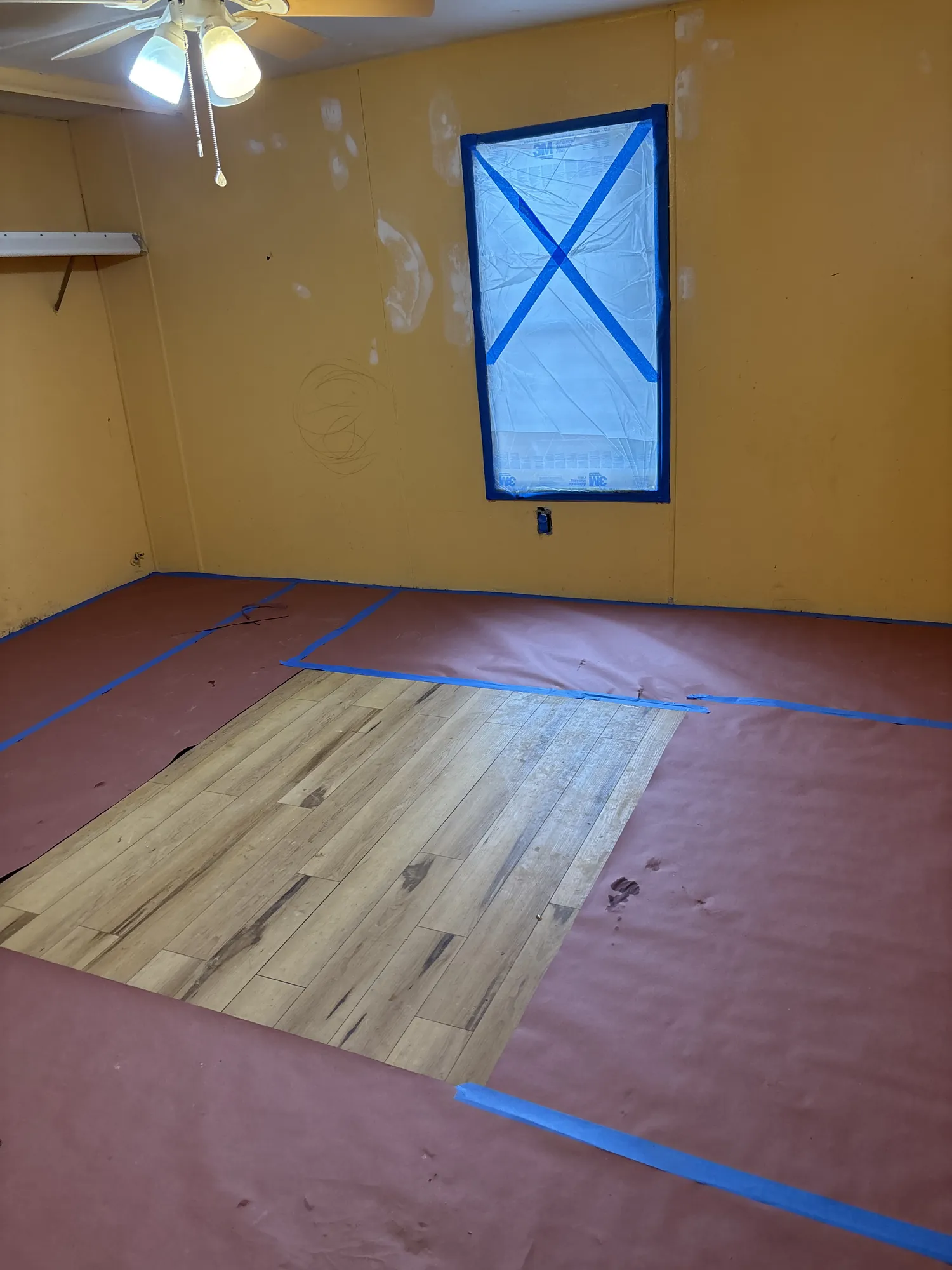 Bedroom with its floor covered in red protective paper and blue tape, a window masked with plastic, and patched orange walls