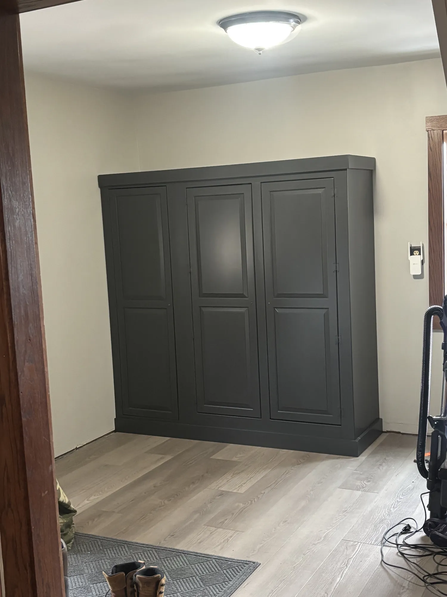 Dark charcoal three-door cabinet with raised panels on gray plank flooring, under a ceiling light, seen through a doorway