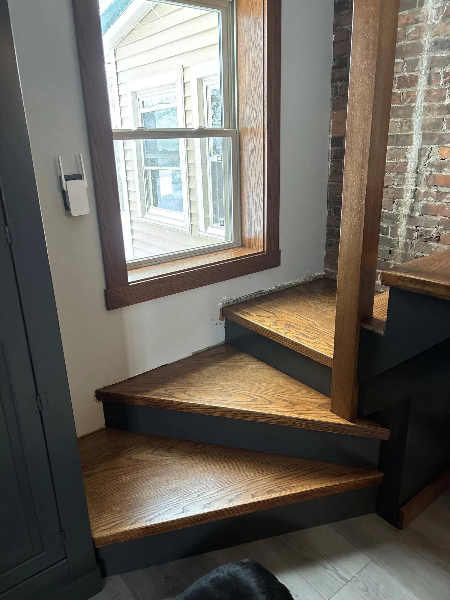 Three stained oak winder steps with charcoal risers turning below a window with stained oak trim, beside exposed brick