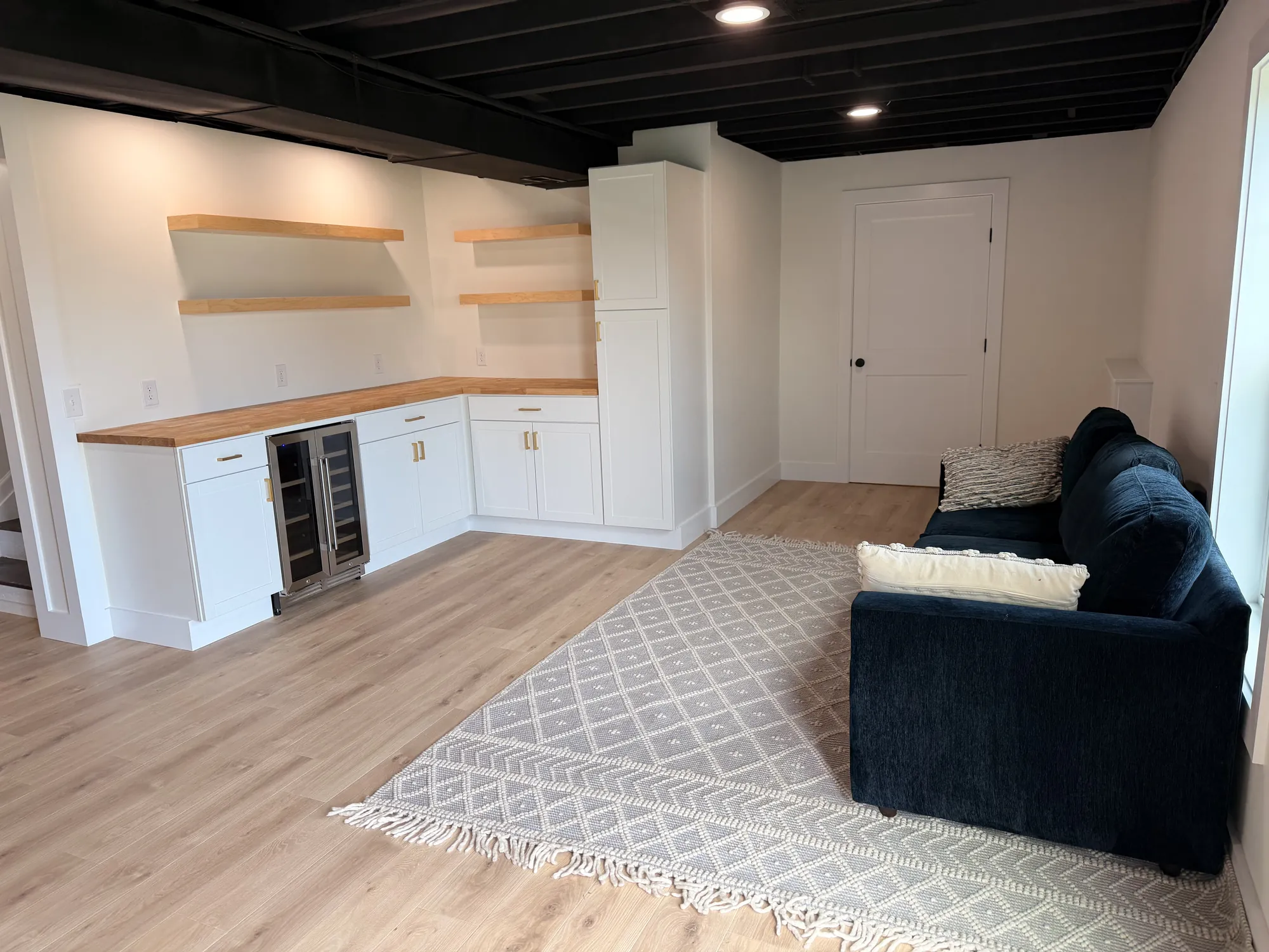 Finished basement with white shaker wet bar, butcher-block counter, floating shelves, beverage fridge, navy sofa and rug