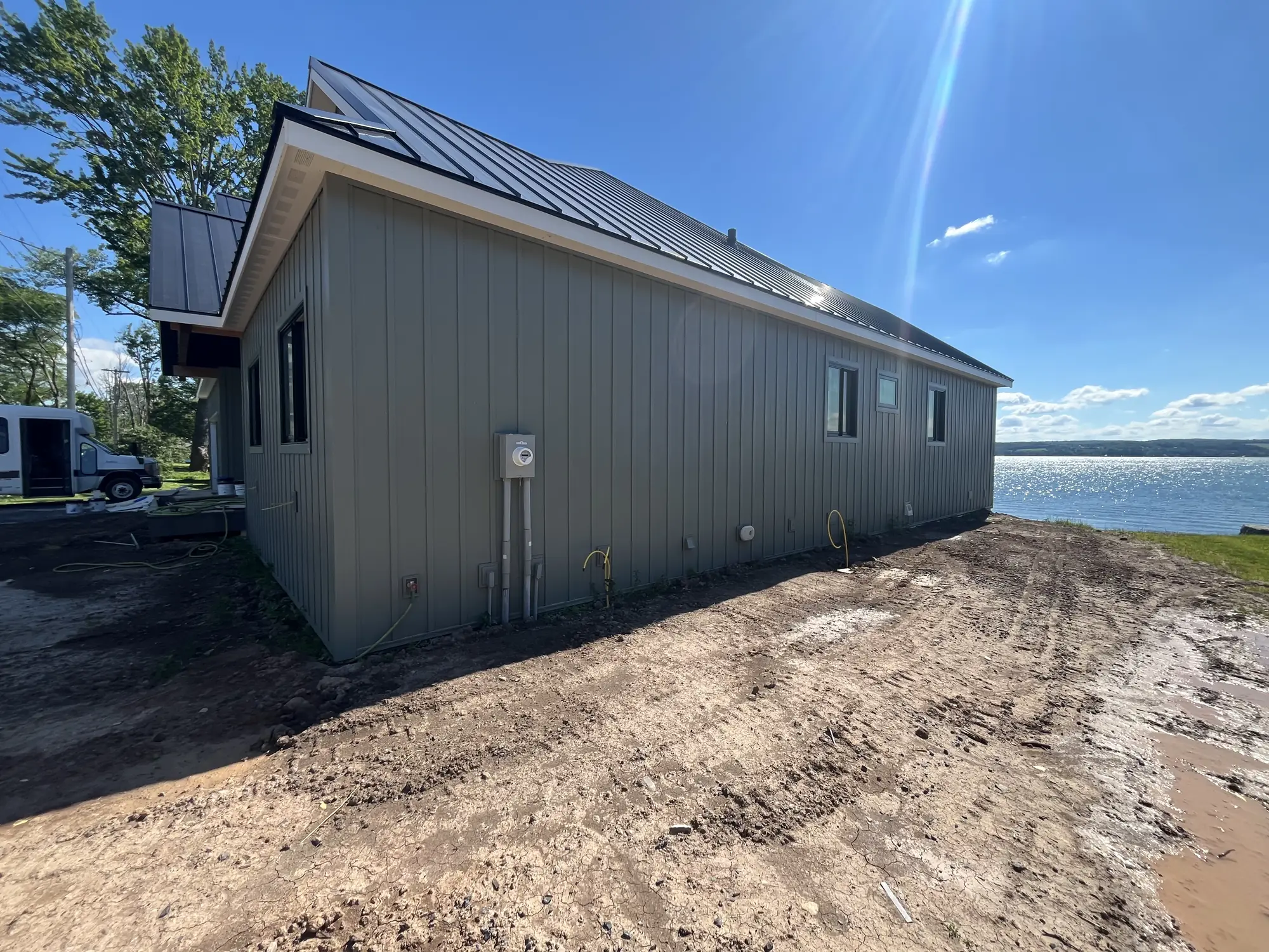 Long board-and-batten side wall under a standing-seam metal roof, with bare dirt in front and sunlit lake water at right