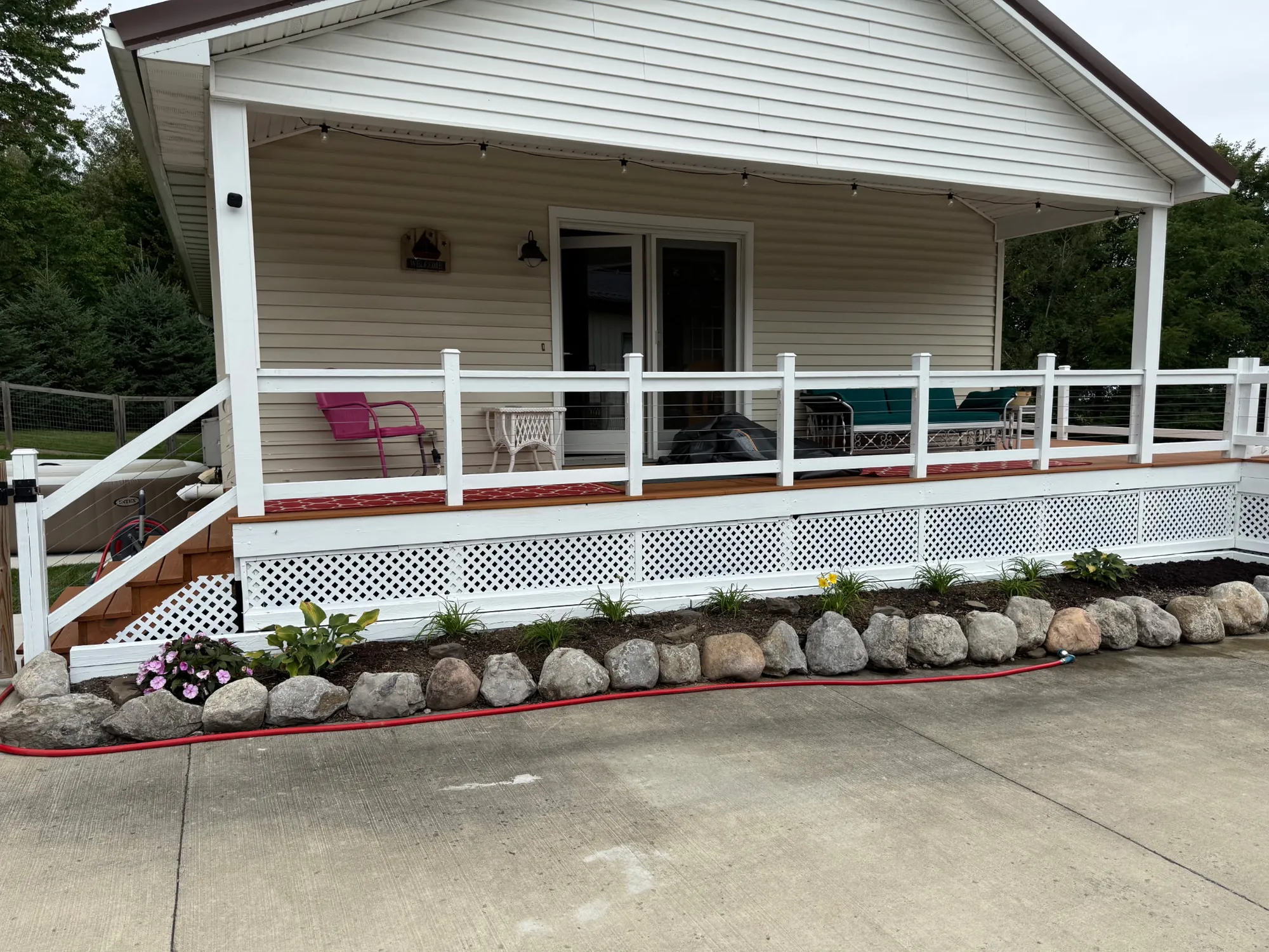 Vinyl-sided cottage with a covered porch, white railings and lattice skirting, stained side steps and a boulder-edged bed