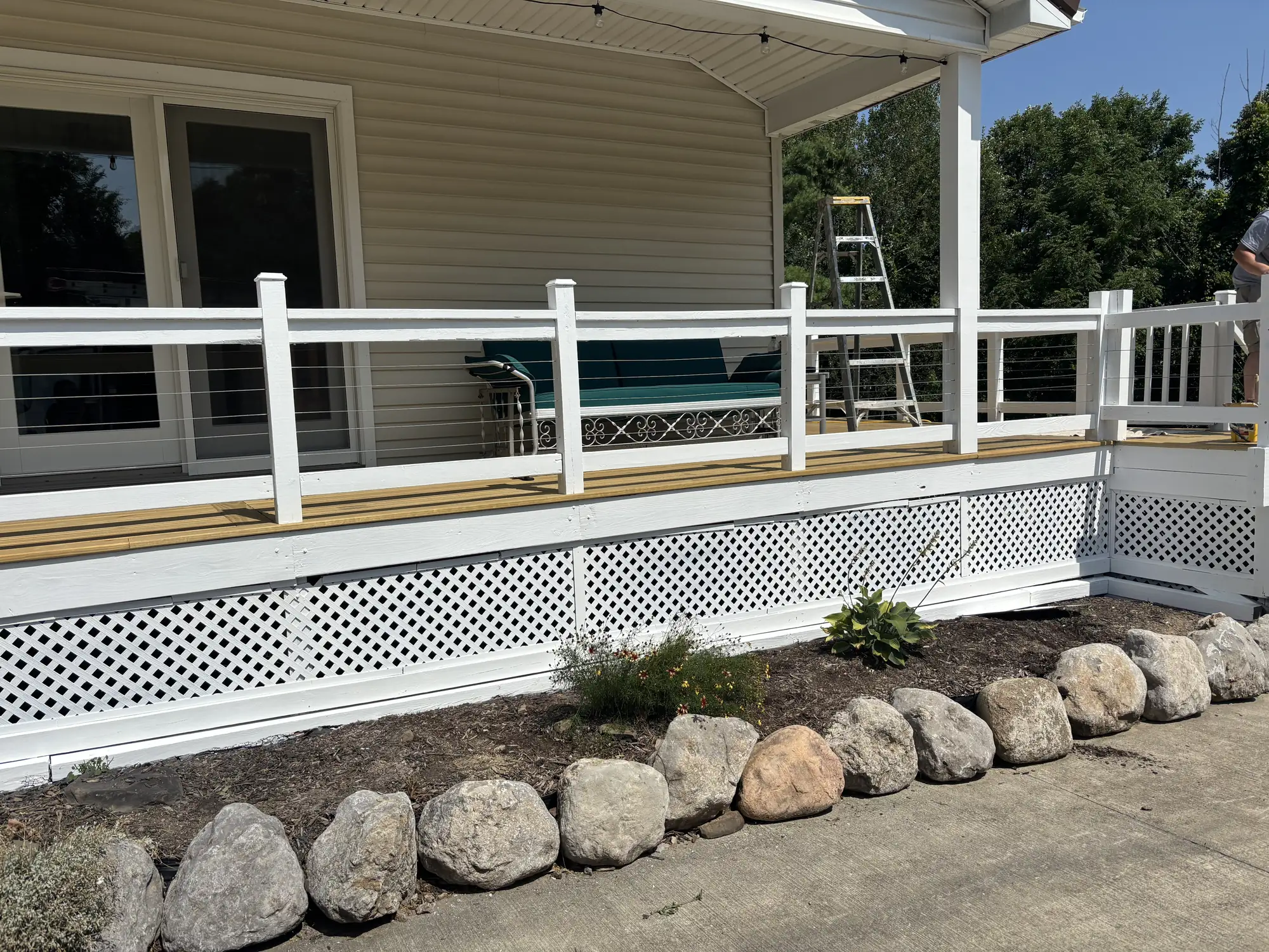 Porch with white railings and cable infill above white lattice skirting, bare deck boards, a stepladder and a boulder edge