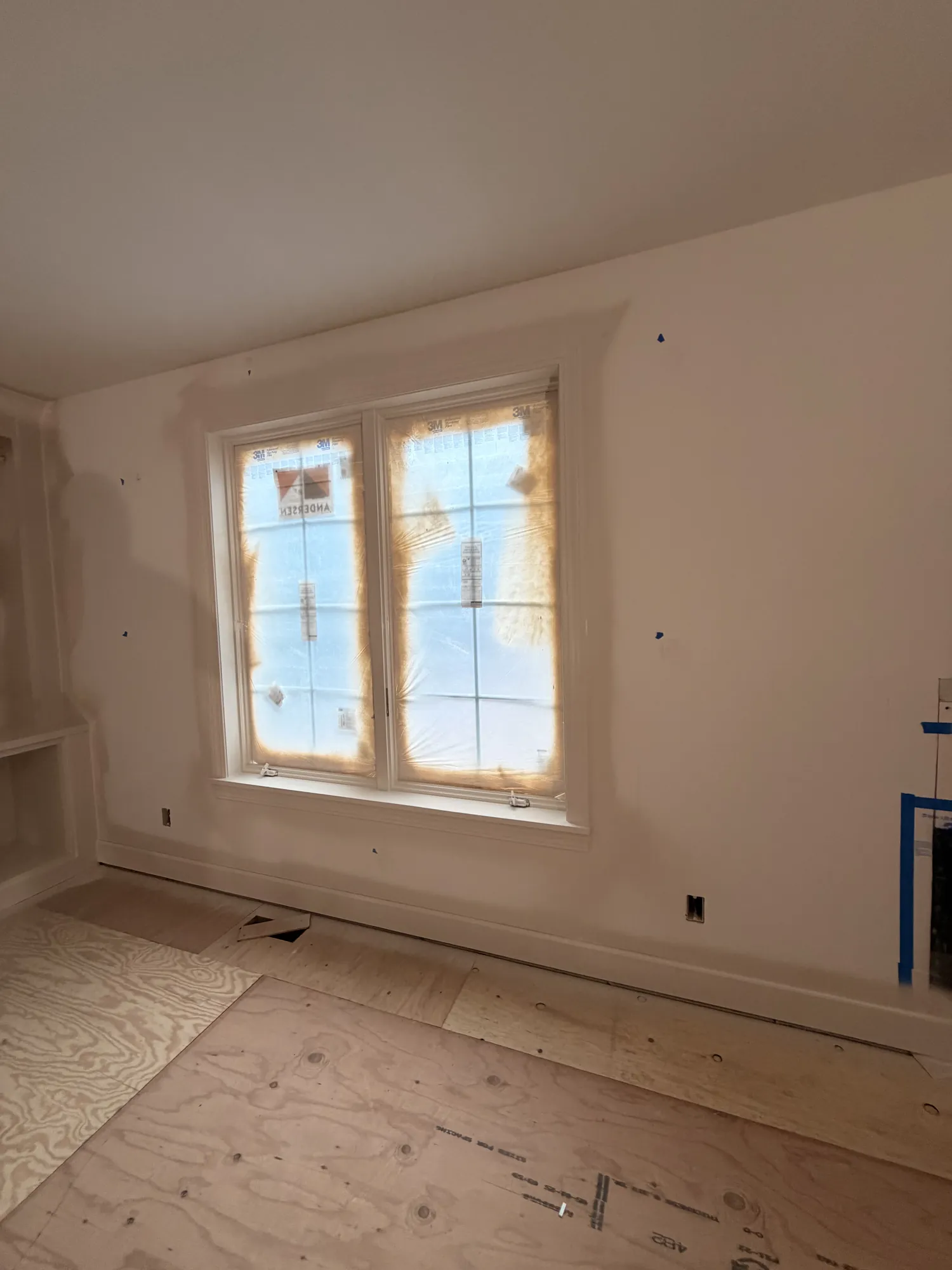 White wall with a double window covered in masking film, a painted window casing, and a bare plywood floor.