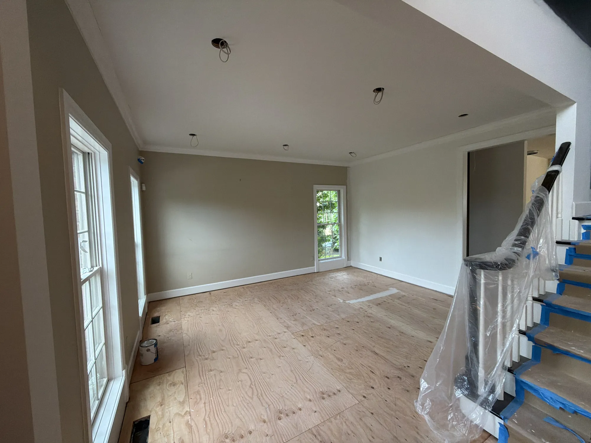 Before: Empty room with beige walls, white trim and crown, tall windows, bare plywood floor, and a stair railing wrapped in plastic.