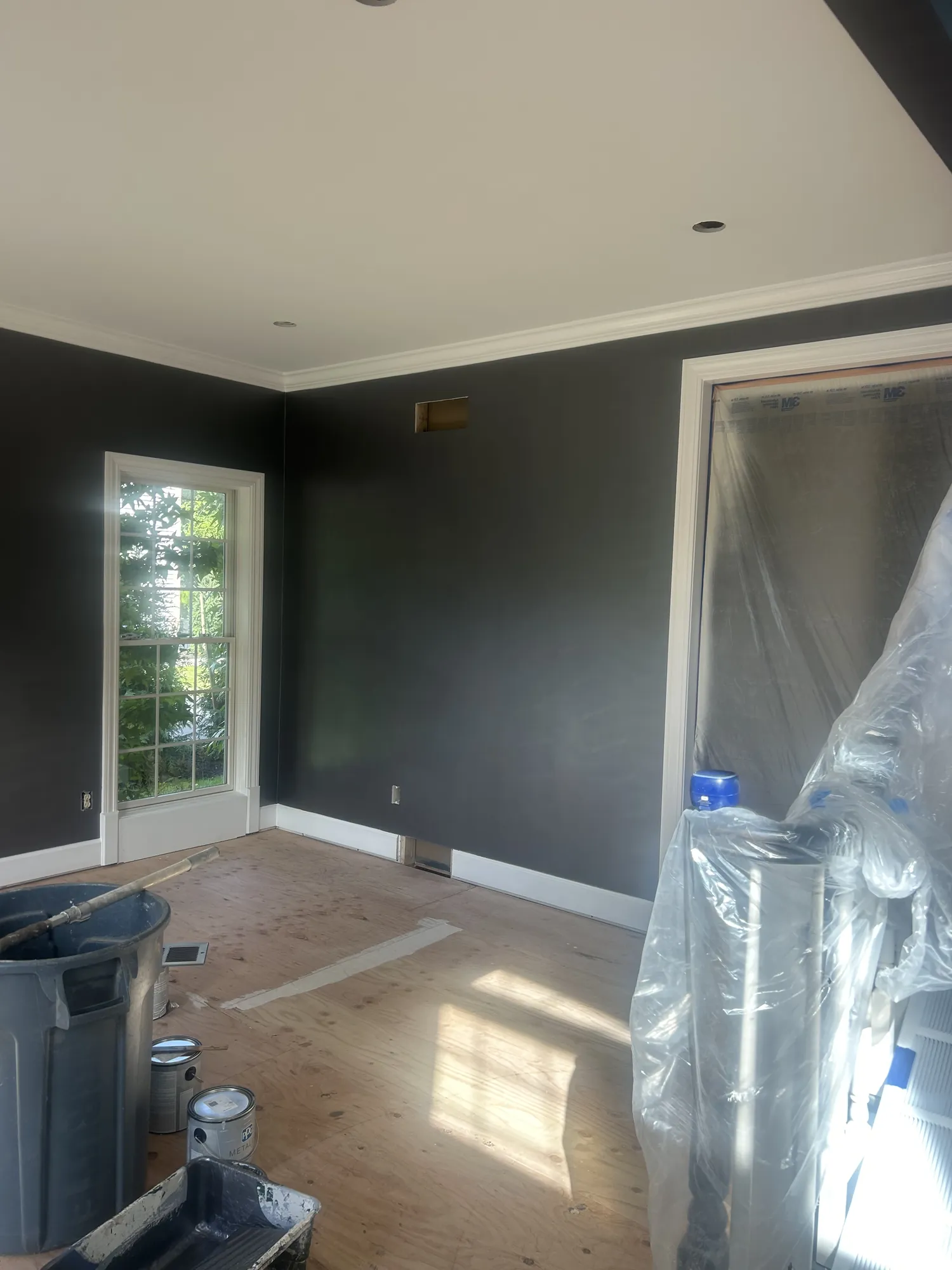 After: Room with deep charcoal walls, white crown and baseboards, a tall window, and paint supplies on a plywood floor.