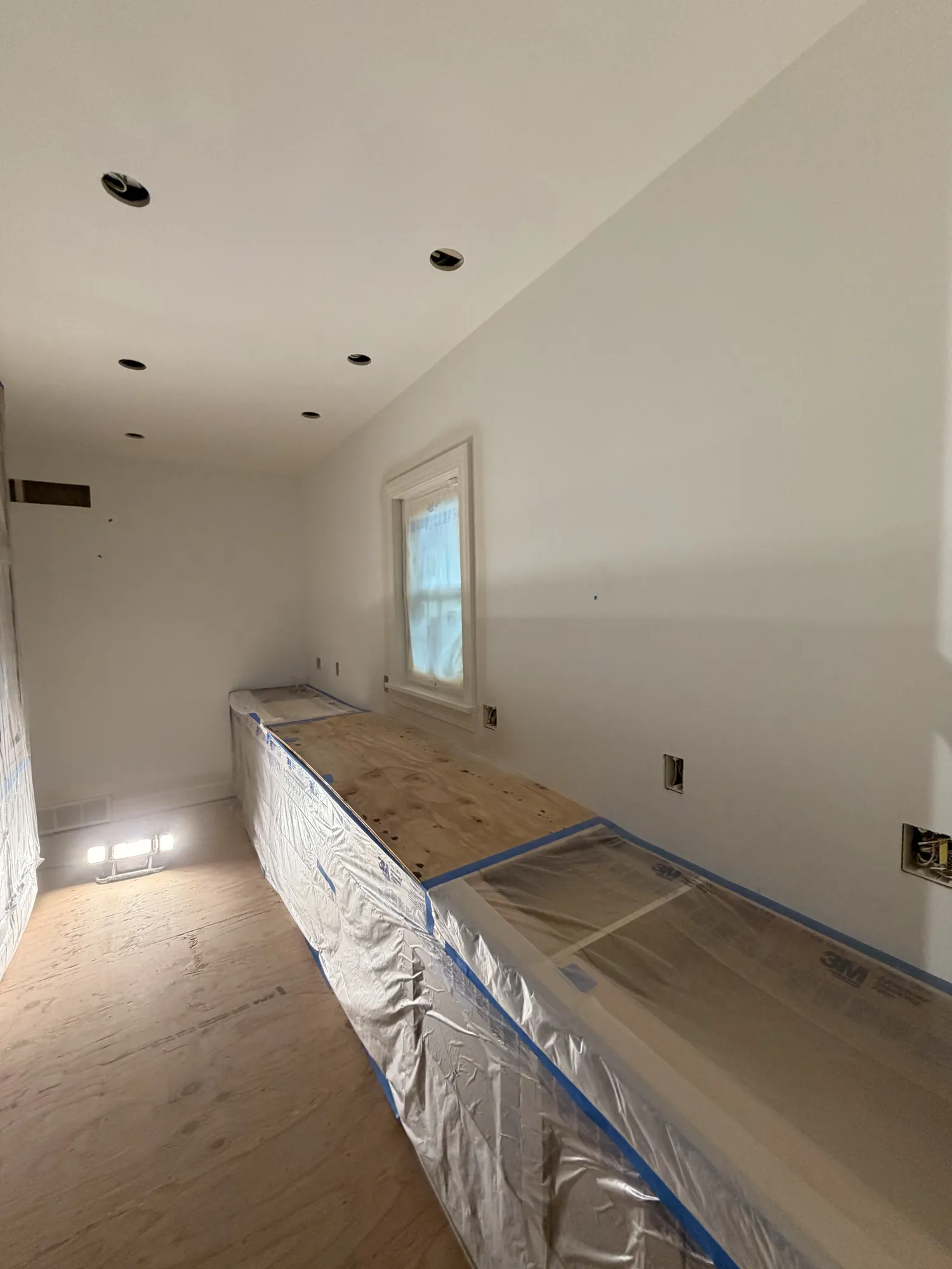 Narrow room with white walls, recessed light openings, and a long counter base wrapped in plastic and masking film.