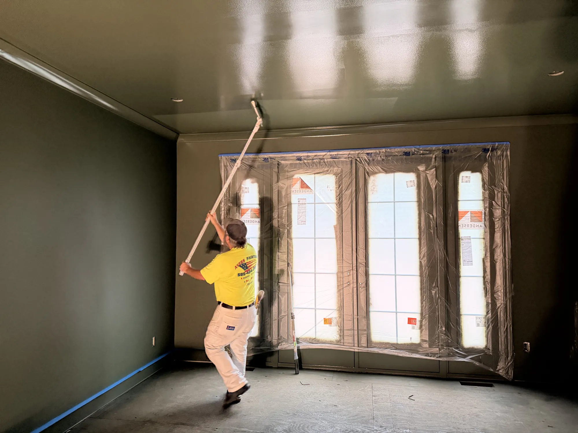 Wide view of a painter in a yellow T-shirt rolling a glossy green ceiling, with deep green walls and masked windows.