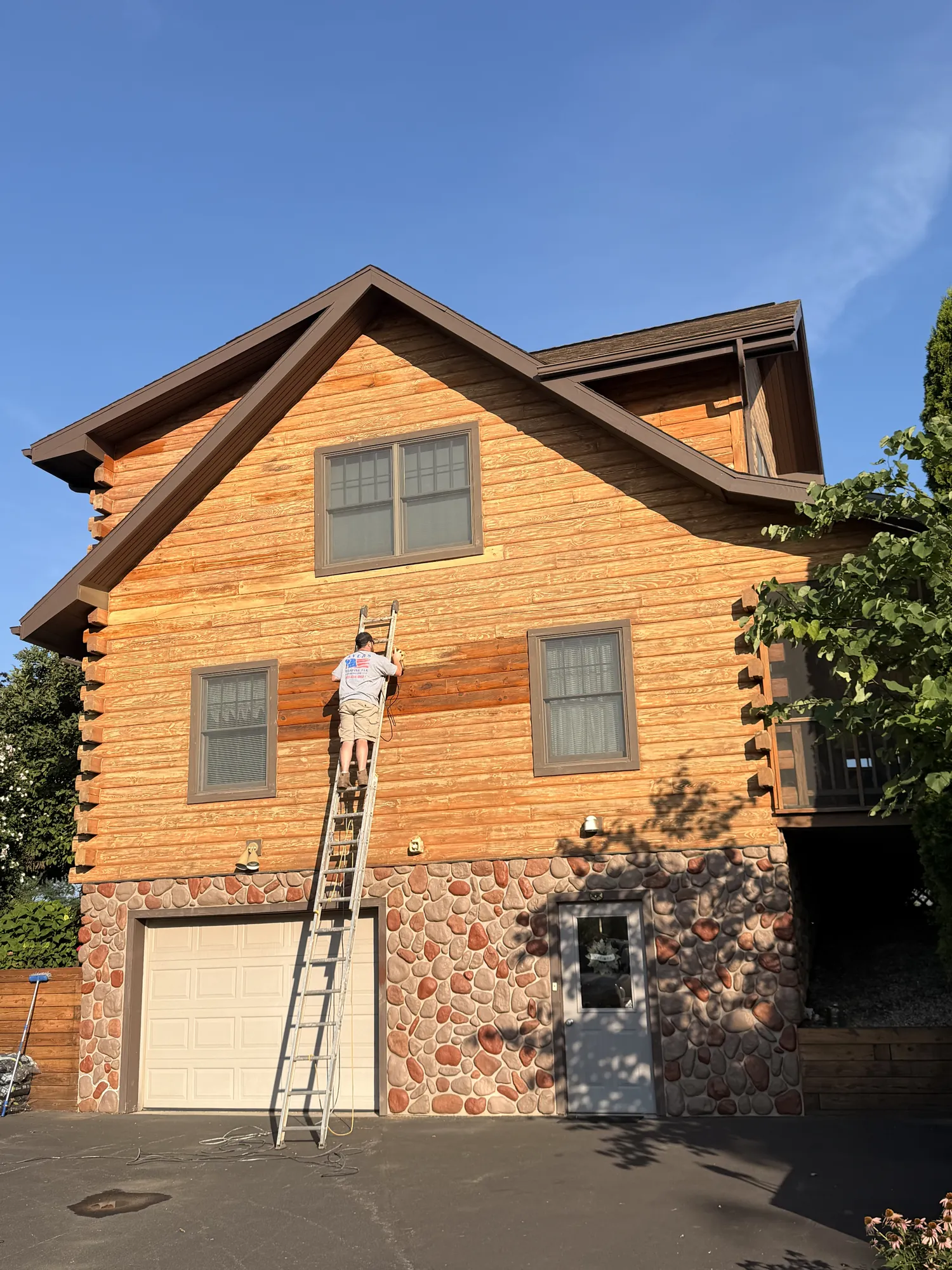 Worker on an extension ladder staining golden log siding, a freshly stained darker band beside him, above a garage with a stone-veneer base