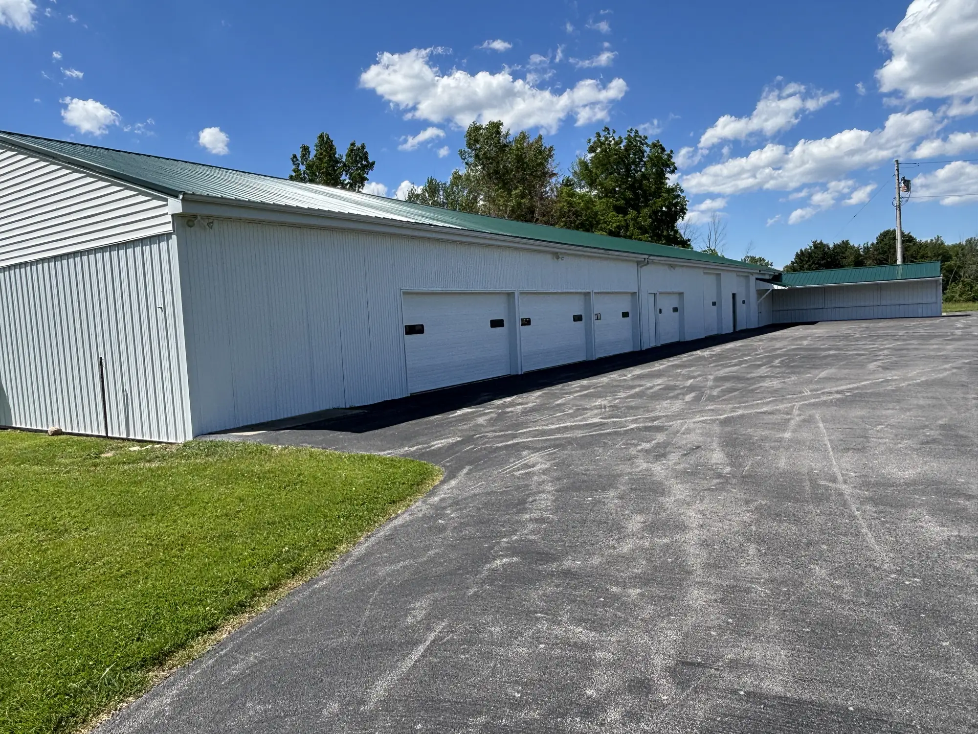 Long white metal building with a green roof and a row of overhead doors beside a fresh asphalt lot under a blue sky.