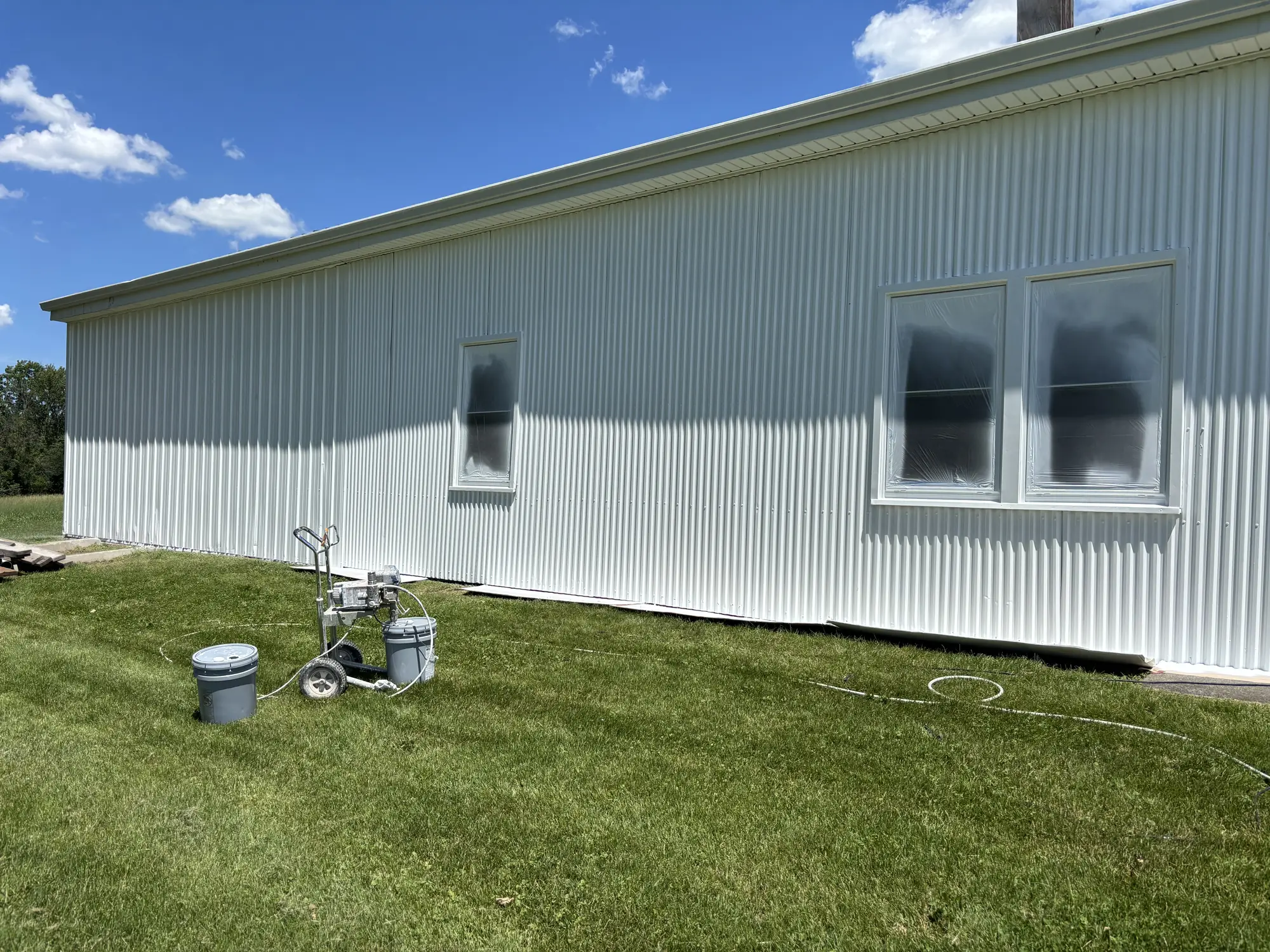 White corrugated metal wall with plastic-covered windows; an airless sprayer and two paint buckets sit on the lawn.
