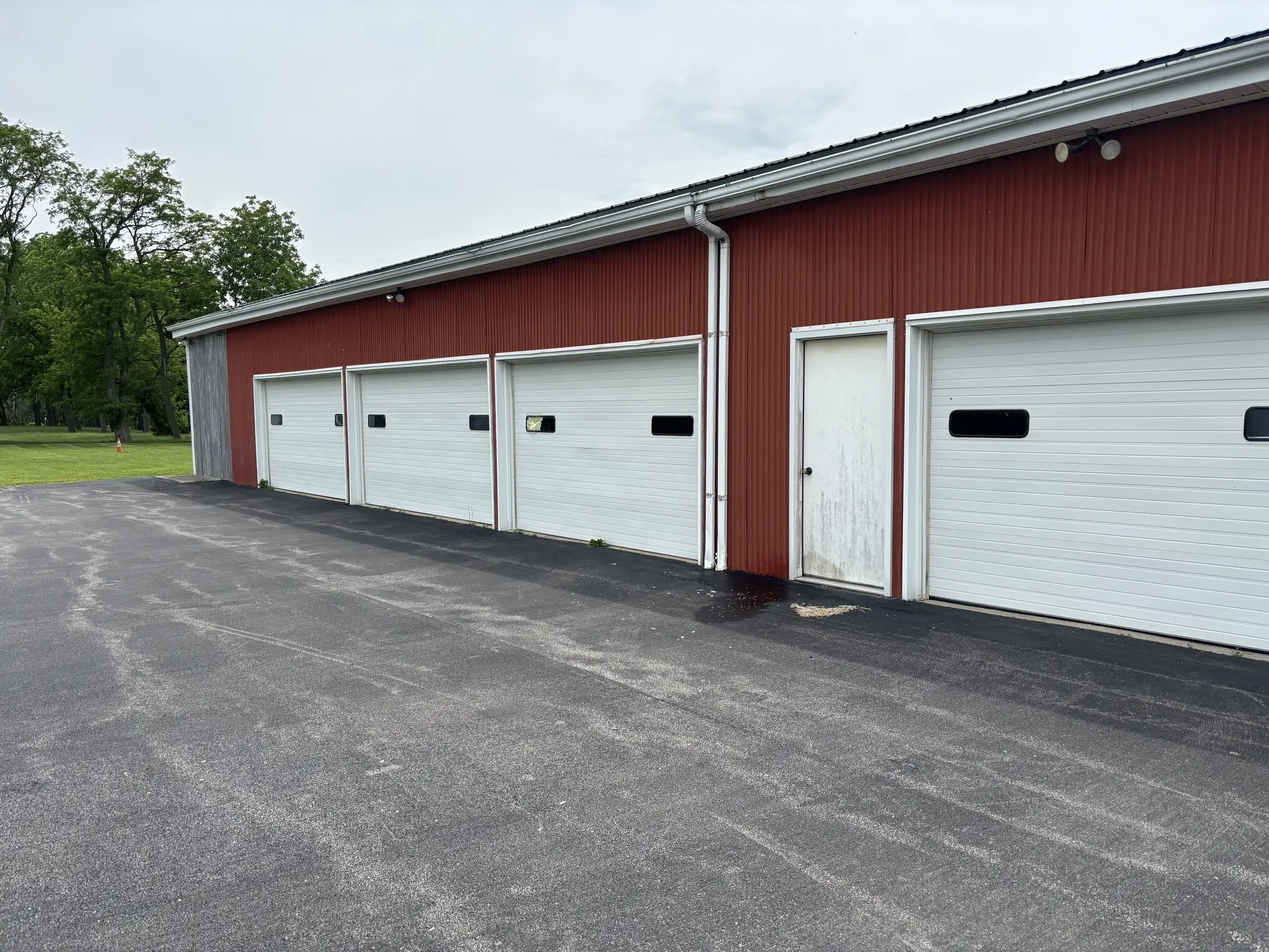 Before: Red corrugated metal wall with four white overhead doors and a stained service door beside an asphalt lot, overcast sky.