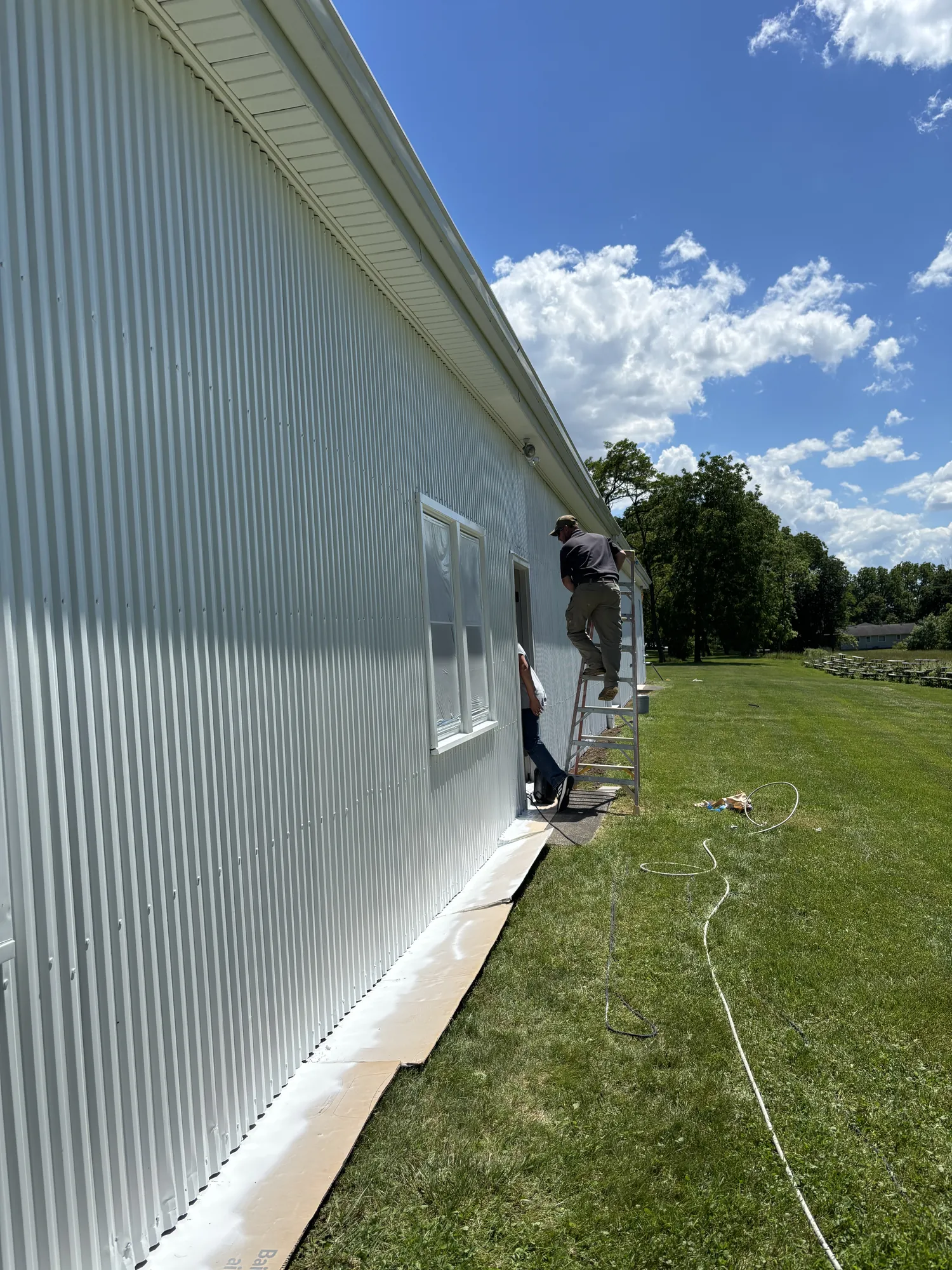 Long white corrugated metal wall with a masked window; one worker on a stepladder and another at the door below, sprayer hose across the lawn