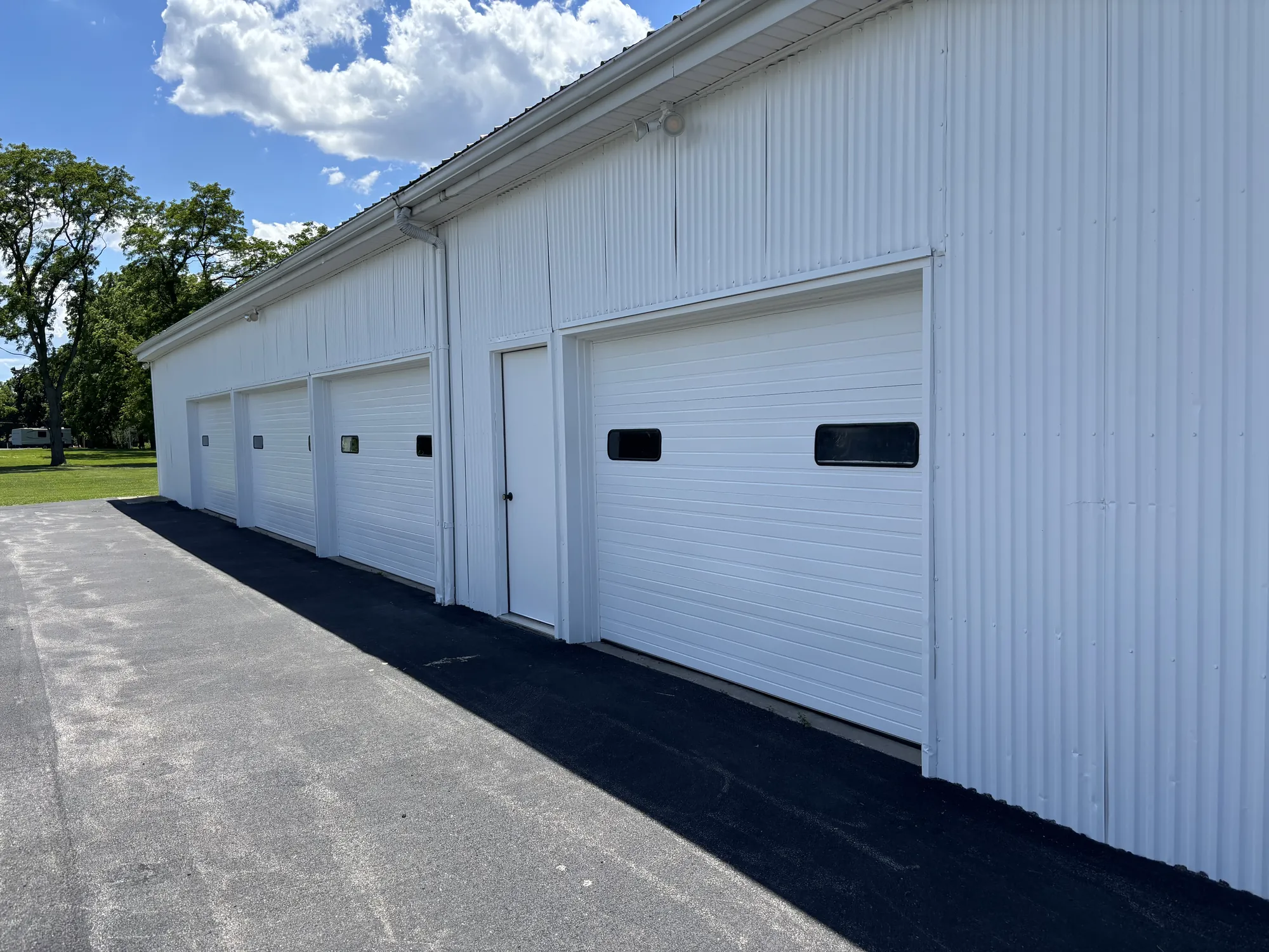 After: Row of white overhead doors along a white corrugated metal wall, fresh asphalt in front, trees and blue sky at left.