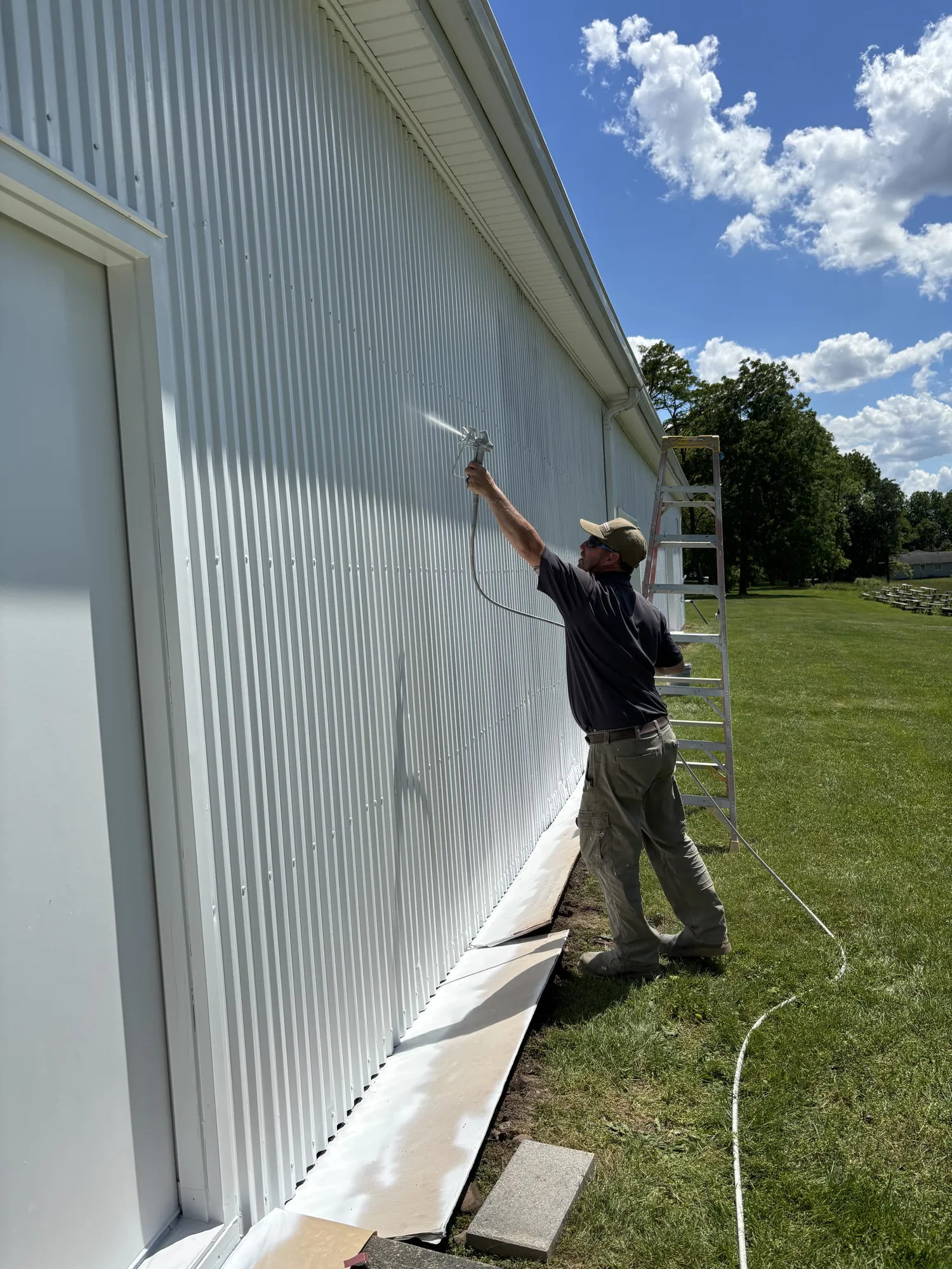 Painter reaches overhead with an airless spray gun, misting white coating onto corrugated metal siding; ladder behind.