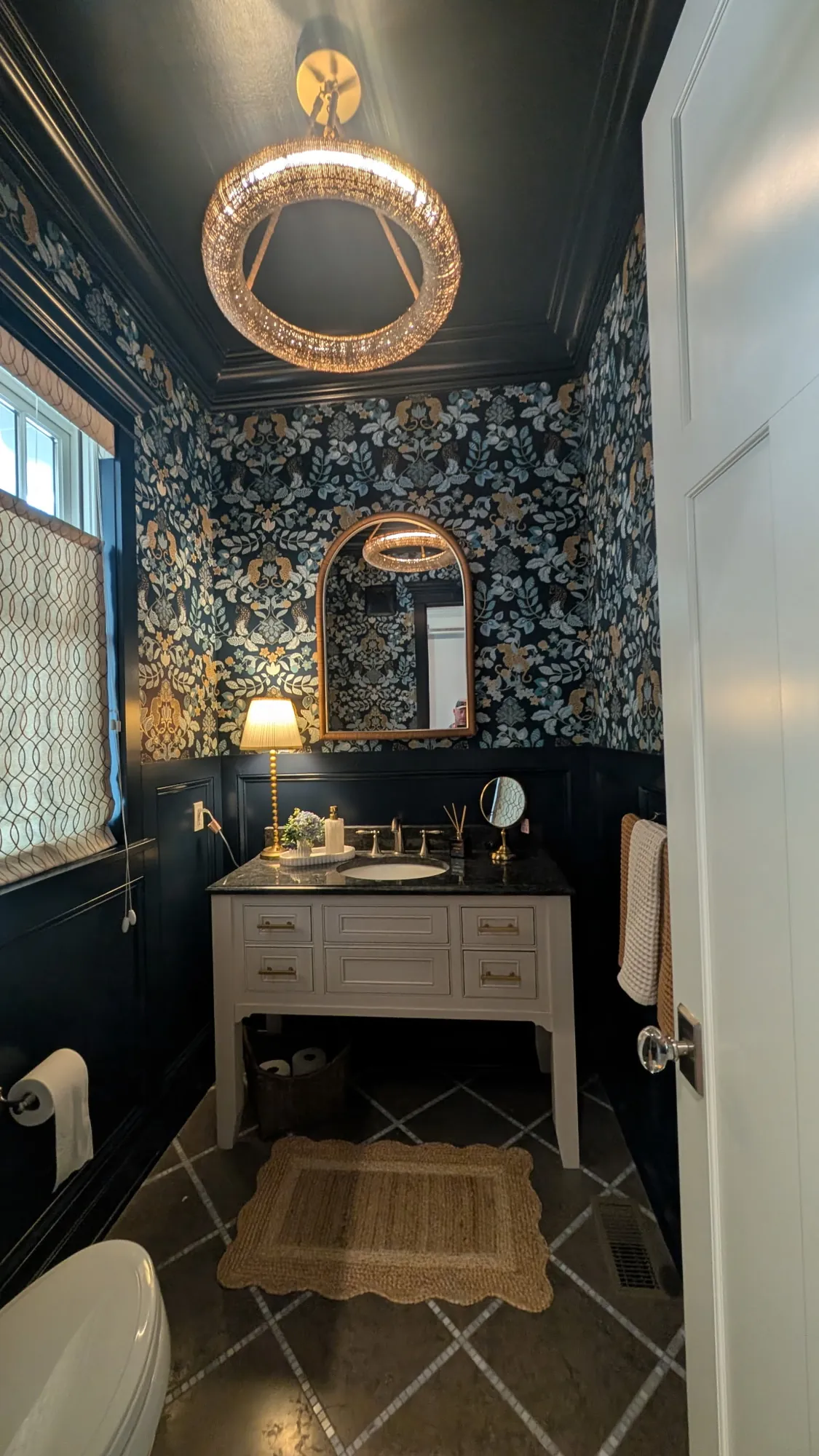 After: Powder room with dark navy wainscoting, crown and ceiling, leaf and leopard wallpaper, a ring chandelier and a white vanity