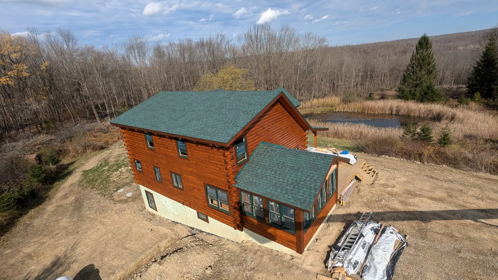 Aerial view of a two-story log cabin with reddish-brown stained logs, a green roof and a sunroom, beside a pond and woods.