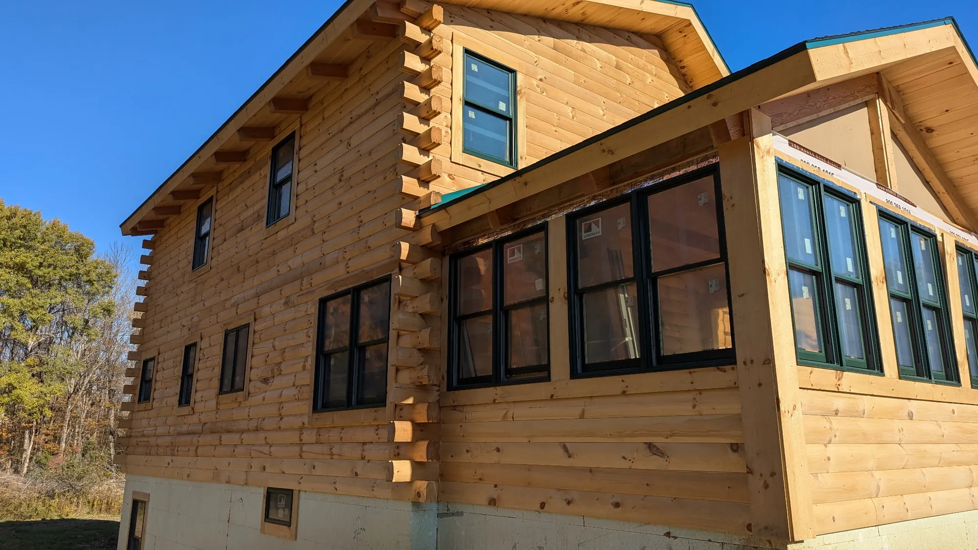 Before: Two-story log cabin in bare, unstained wood with dark-framed windows and a sunroom, under a clear blue sky.