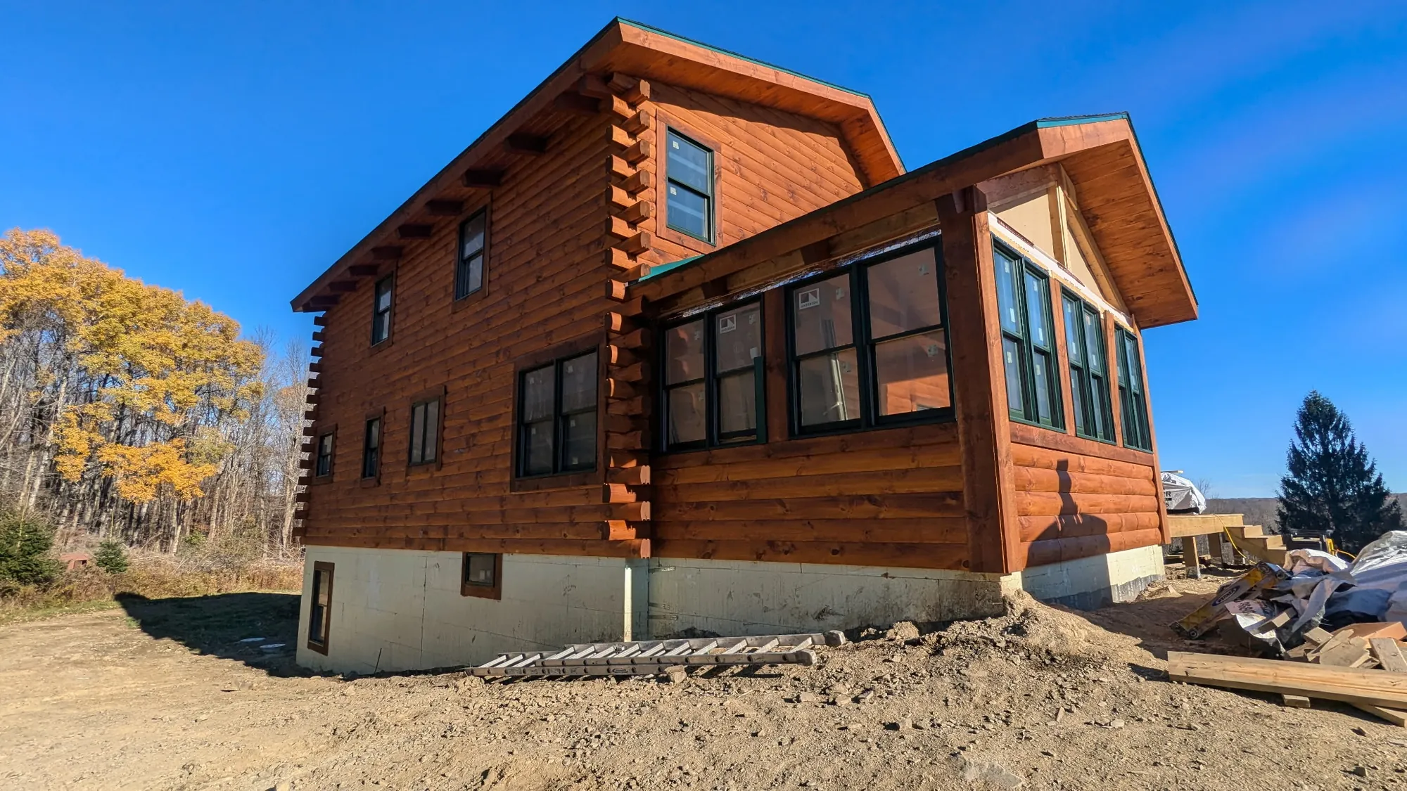 After: Two-story log cabin with freshly stained reddish-brown logs, dark-framed windows and a sunroom, under a clear blue sky.