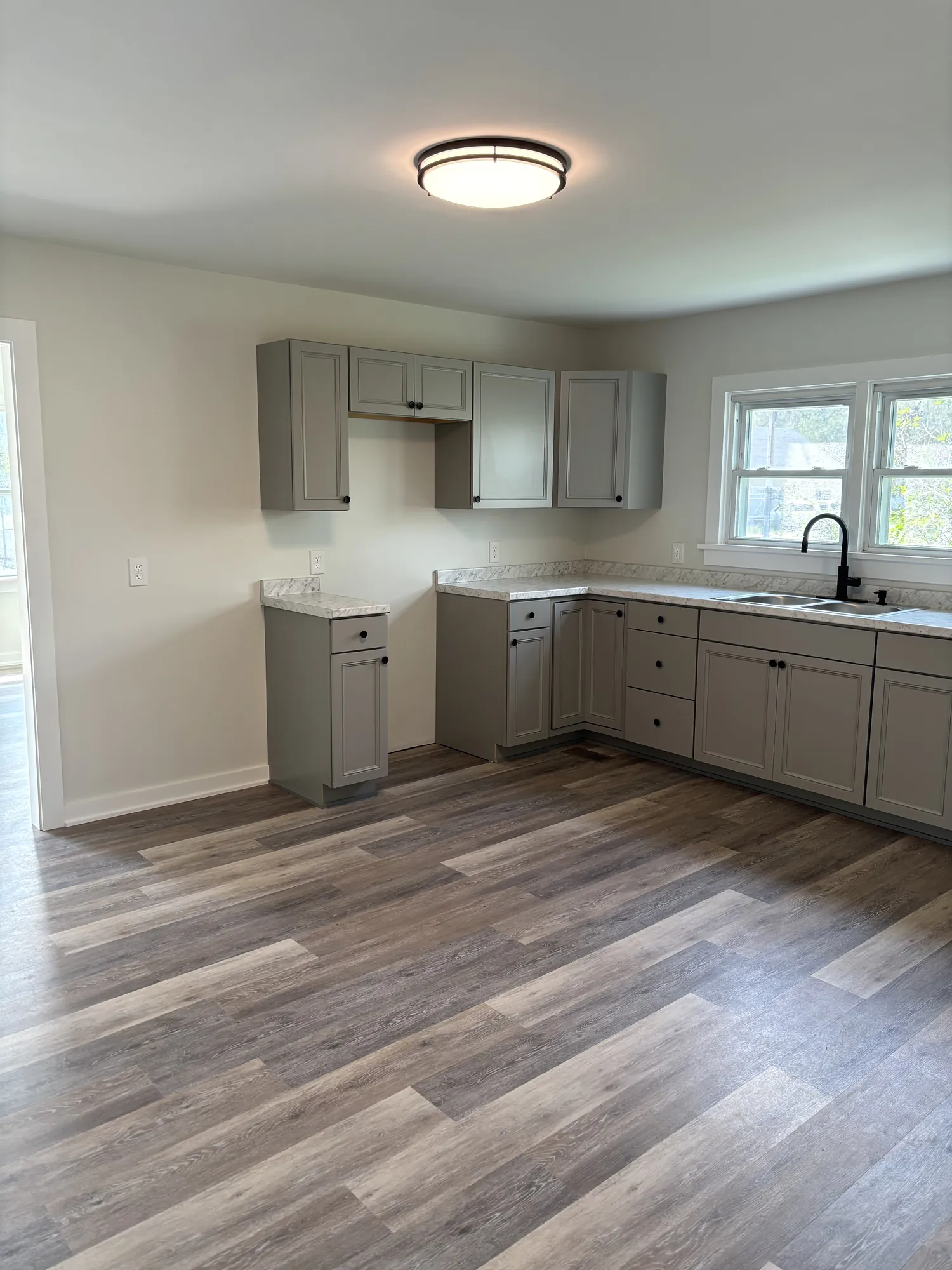 After: Kitchen with gray shaker-style cabinets, stone-look counters, black faucet under a double window, plank floor, ceiling light