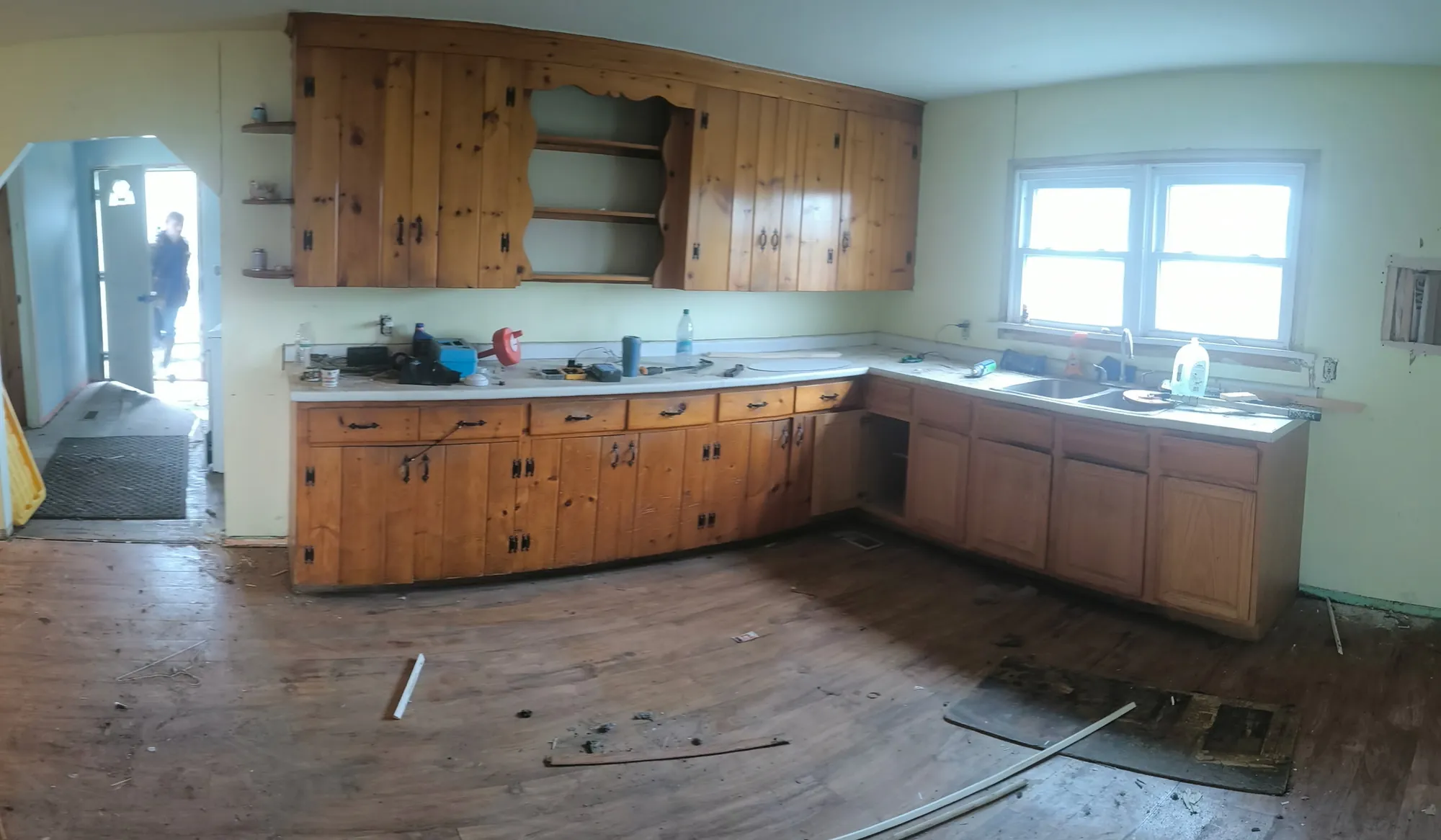 Before: Dated kitchen with knotty pine upper and lower cabinets in an L shape, a window over the sink and a worn, damaged wood floor