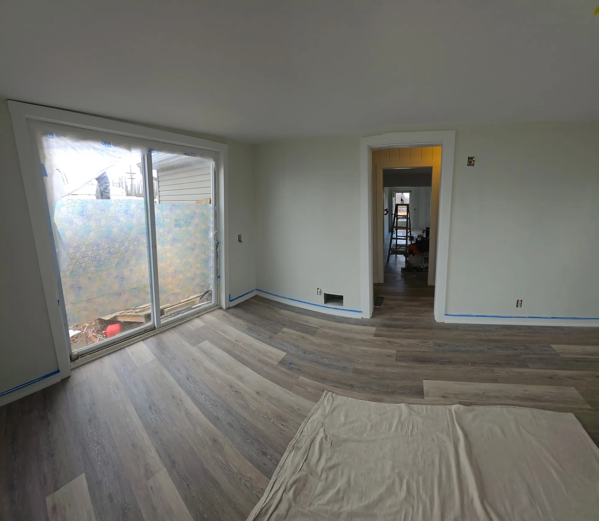 Room with a sliding door masked in plastic, blue tape on the baseboards, a drop cloth on new plank flooring, a ladder beyond