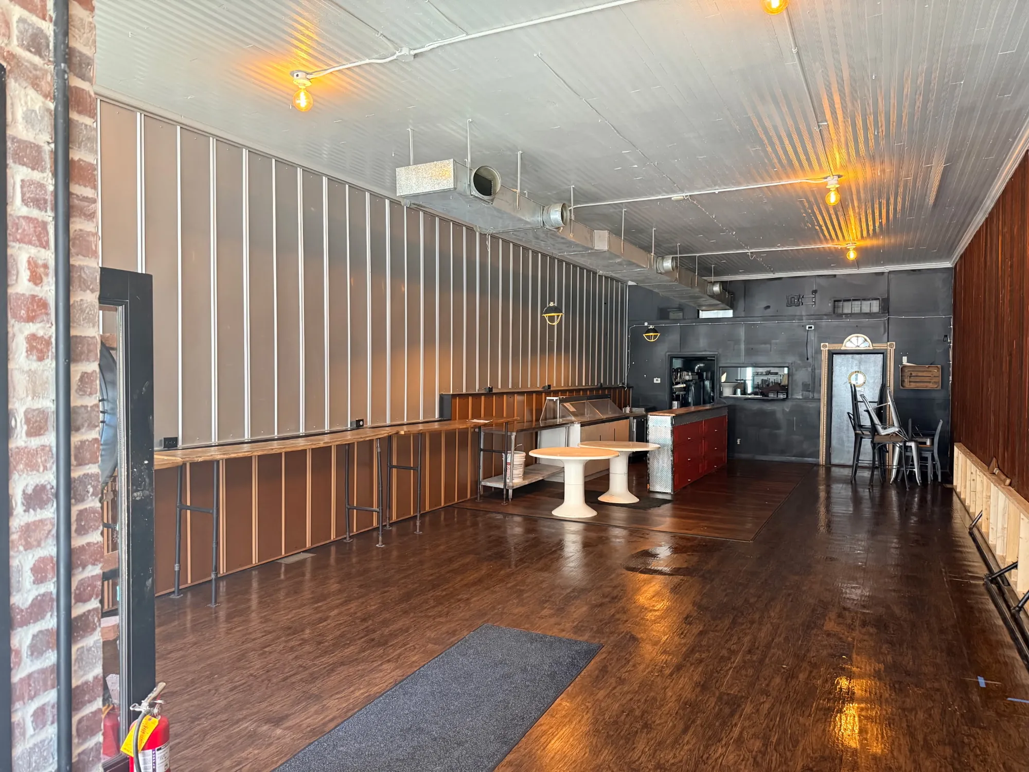 Long restaurant dining room with its ribbed ceiling sprayed a light fire-retardant coating, exposed ductwork, a metal-panel wall, a counter and a dark plank floor