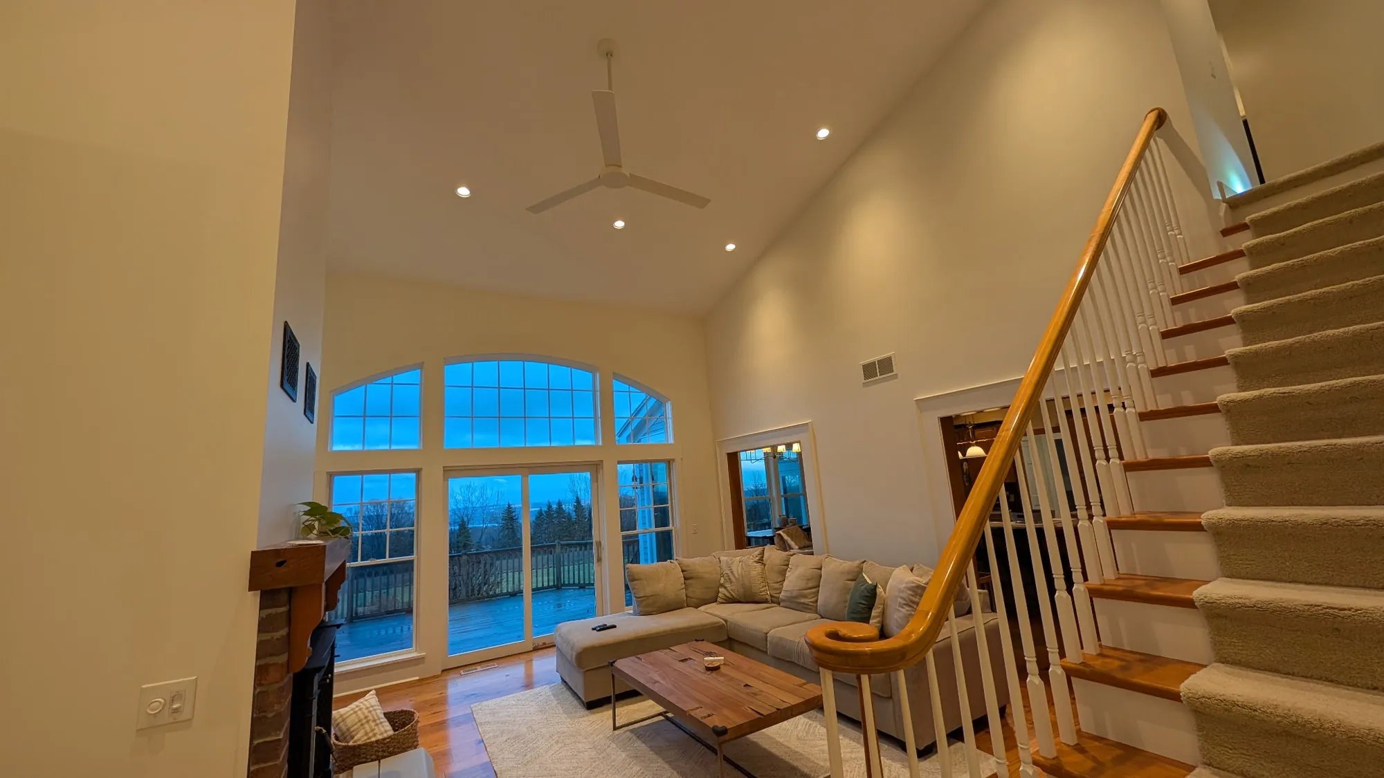 After: Vaulted living room at dusk with recessed lights, white-trimmed arched windows and a staircase with white balusters